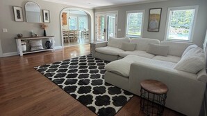 Uma sala de estar ampla com assoalho de madeira, um sofá seccional grande, um tapete preto e branco e uma área de jantar ao fundo.