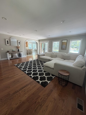 A spacious living room with hardwood flooring, a large sectional sofa, a black and white rug, and a dining area in the background.