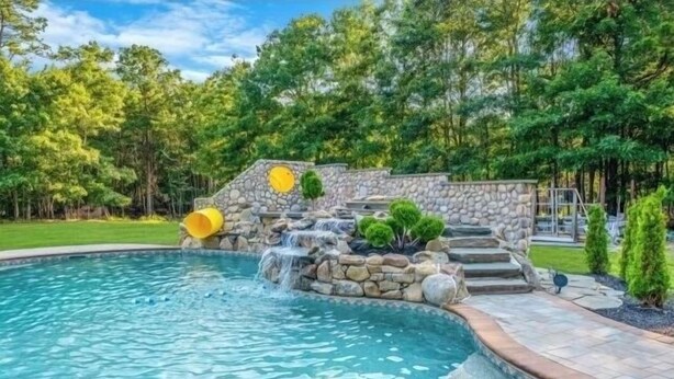 Uma piscina com uma cascata de pedra e degraus que a levam, rodeada por vegetação exuberante.