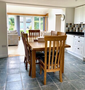 A dining area with a wooden table and chairs, a kitchen with white cabinets, and a view of greenery through large windows.