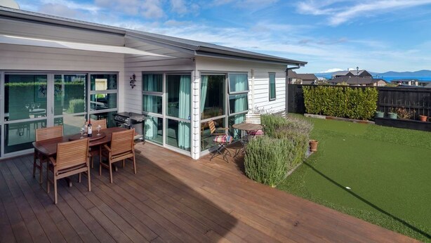 Une maison moderne dotée d’une terrasse en bois, d’un patio fermé par des baies vitrées et d’une pelouse bien entretenue.