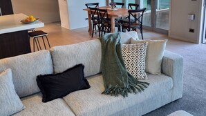 Un salon moderne avec un canapé d’angle gris, un coin repas comprenant une table et des chaises en bois, et une grande fenêtre laissant entrer la lumière naturelle.