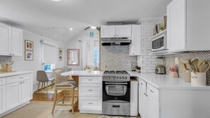 A modern kitchen with white cabinetry, a built-in oven, and a dining area with a chair and table. - The Daydreamer - Walk To Walnut Beach, Restaurant + Shops (Milford)