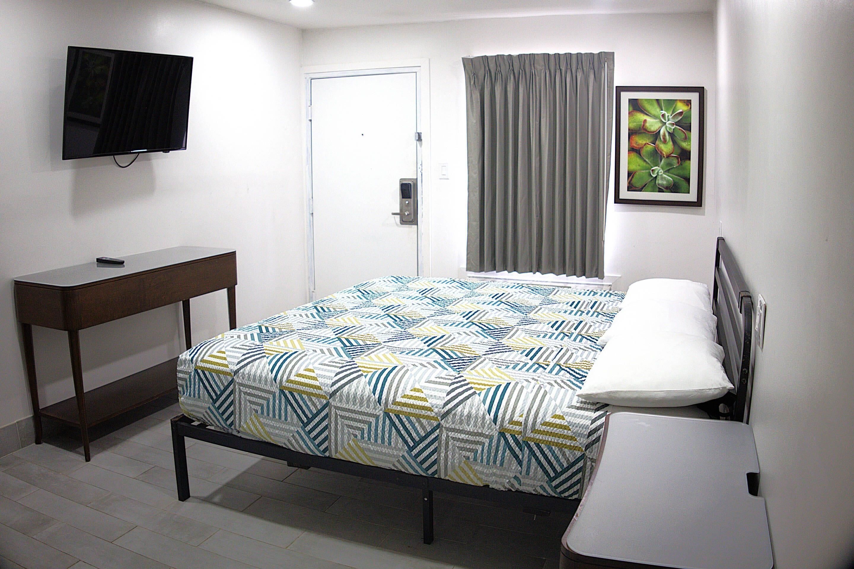 Ein Hotelzimmer mit Bett, Schreibtisch, Wandfernseher, Tür und einem fensterbepflanzten Fenster.