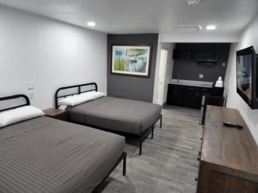 A room with two beds, a wooden desk, a TV, and a kitchen area.