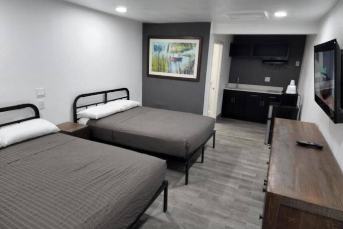 A room with two beds, a wooden desk, a TV, and a kitchen area.