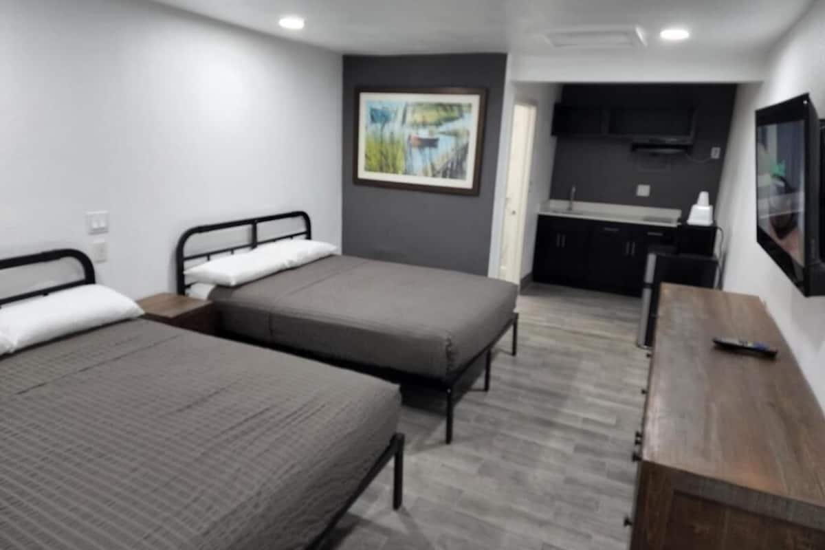 A room with two beds, a wooden desk, a TV, and a kitchen area.