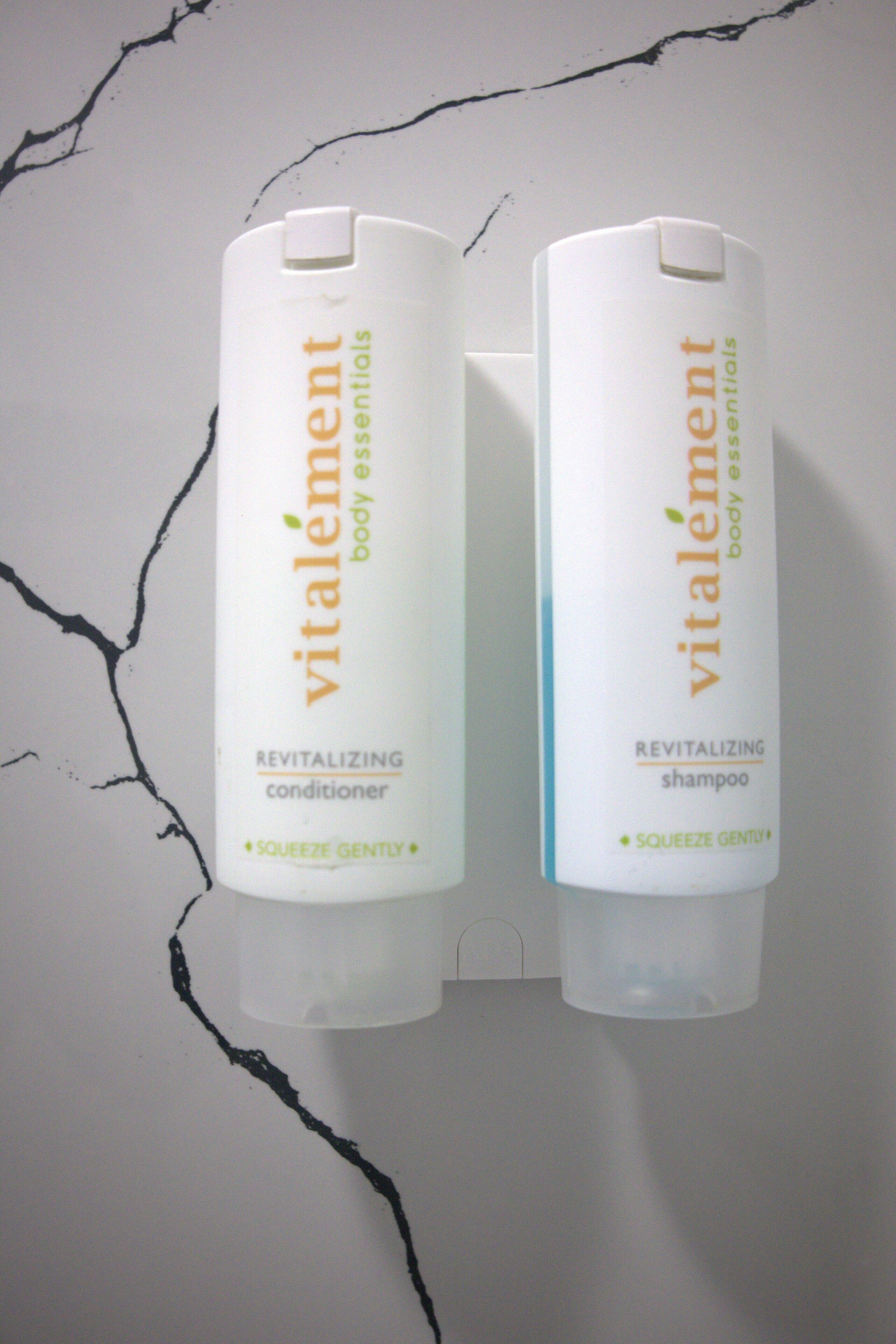 Two bottles of Vitalement body essentials, one conditioner and one shampoo, with a cracked wall in the background.