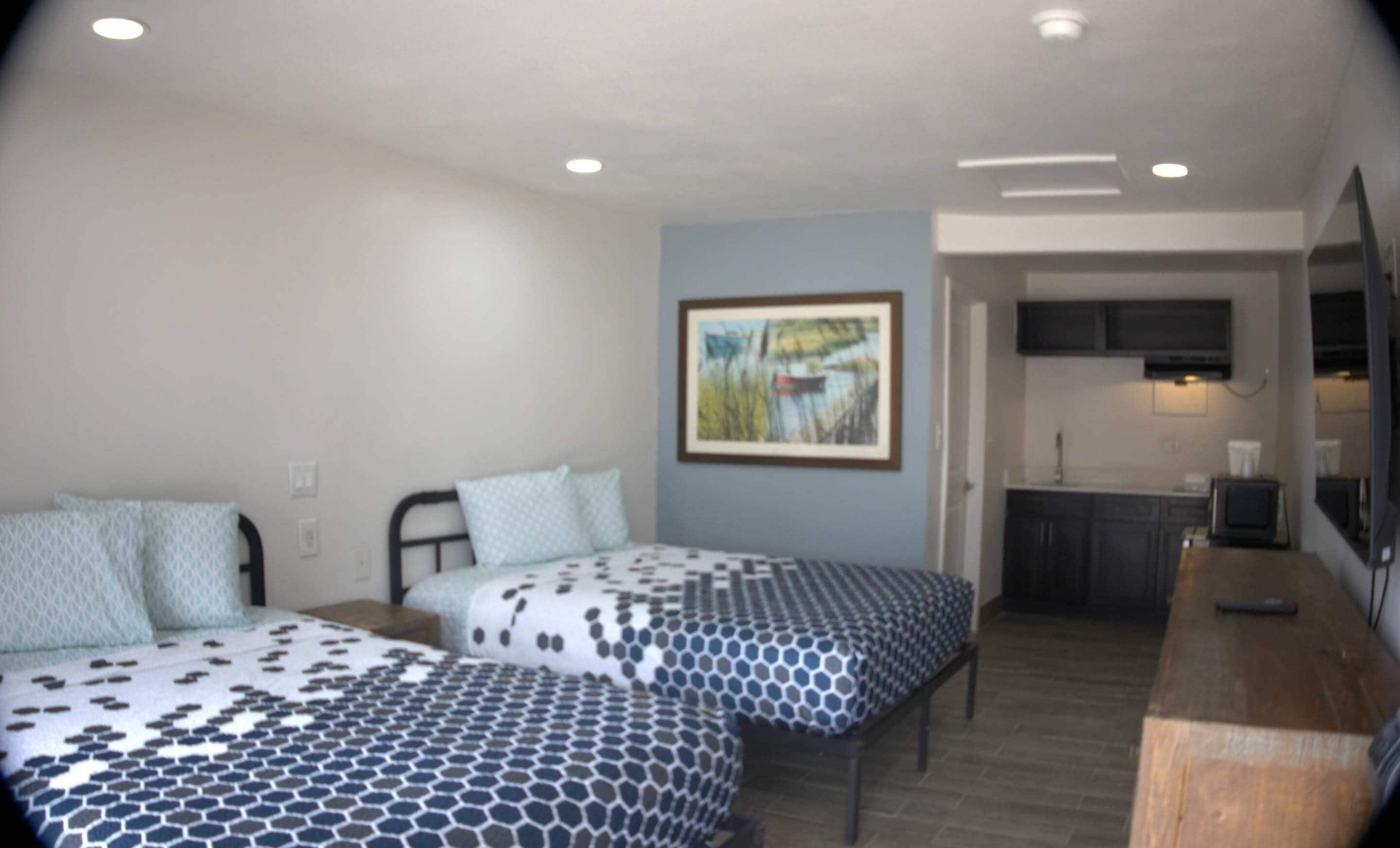 A room with two beds, a painting on the wall, and a kitchenette in the background.
