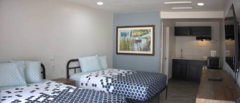 A room with two beds, a painting on the wall, and a kitchenette in the background.