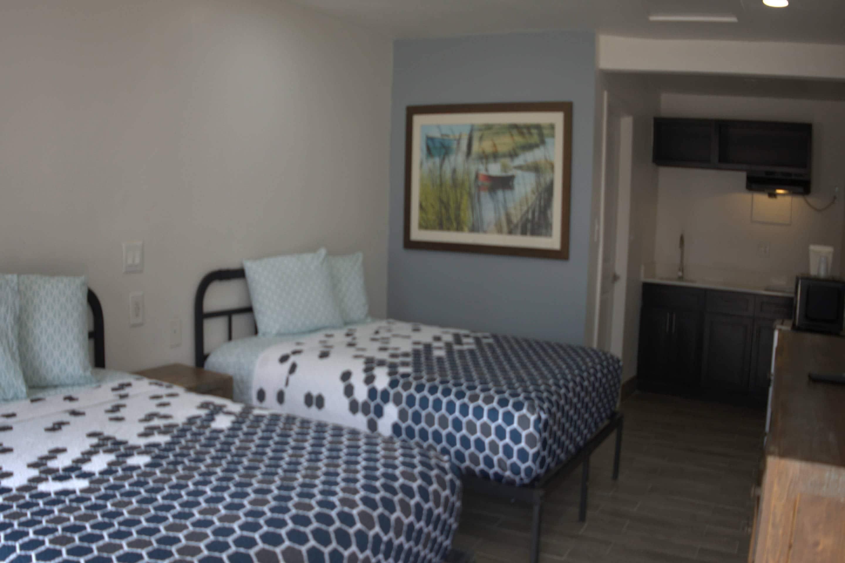 A bedroom with two beds, a painting on the wall, and a kitchenette in the background.