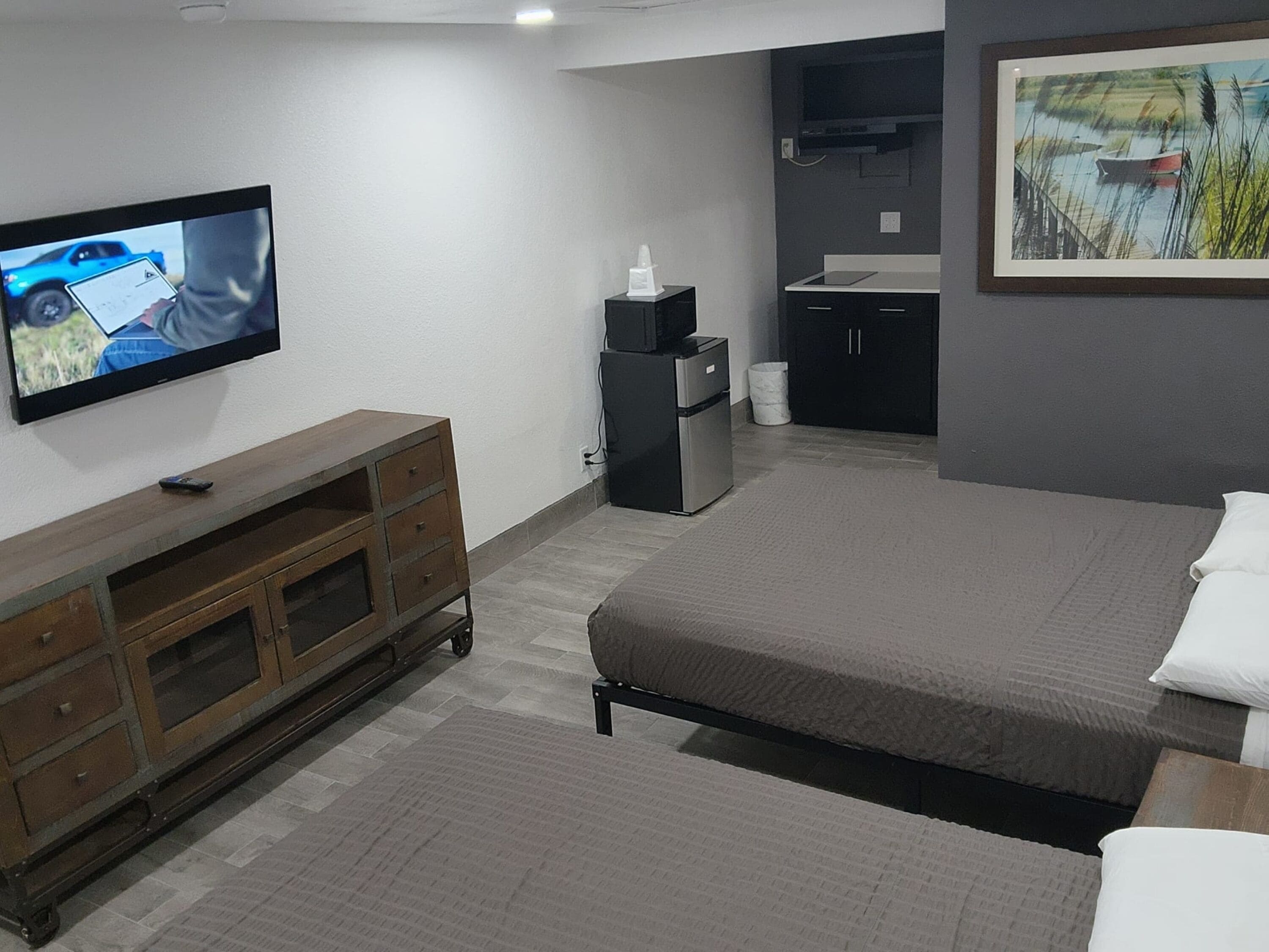 Ein Hotelzimmer mit einem Bett, einem an der Wand montierten Fernseher, einer Holzkommode, einem Kühlschrank und einem Bild an der Wand.
