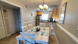 A dining area with a white table set for six, surrounded by white chairs. A chandelier hangs from the ceiling, and a kitchen is visible in the background. - 2 minutes to the beach (Pensacola Beach)