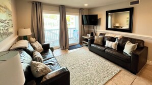 A living room with a black leather sofa, a matching armchair, a large flat-screen TV mounted on the wall, and a sliding glass door leading to an outdoor area. - 2 minutes to the beach (Pensacola Beach)