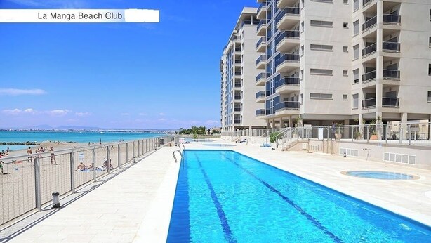 Une piscine située à côté d’une plage, avec un immeuble à plusieurs étages en arrière-plan.