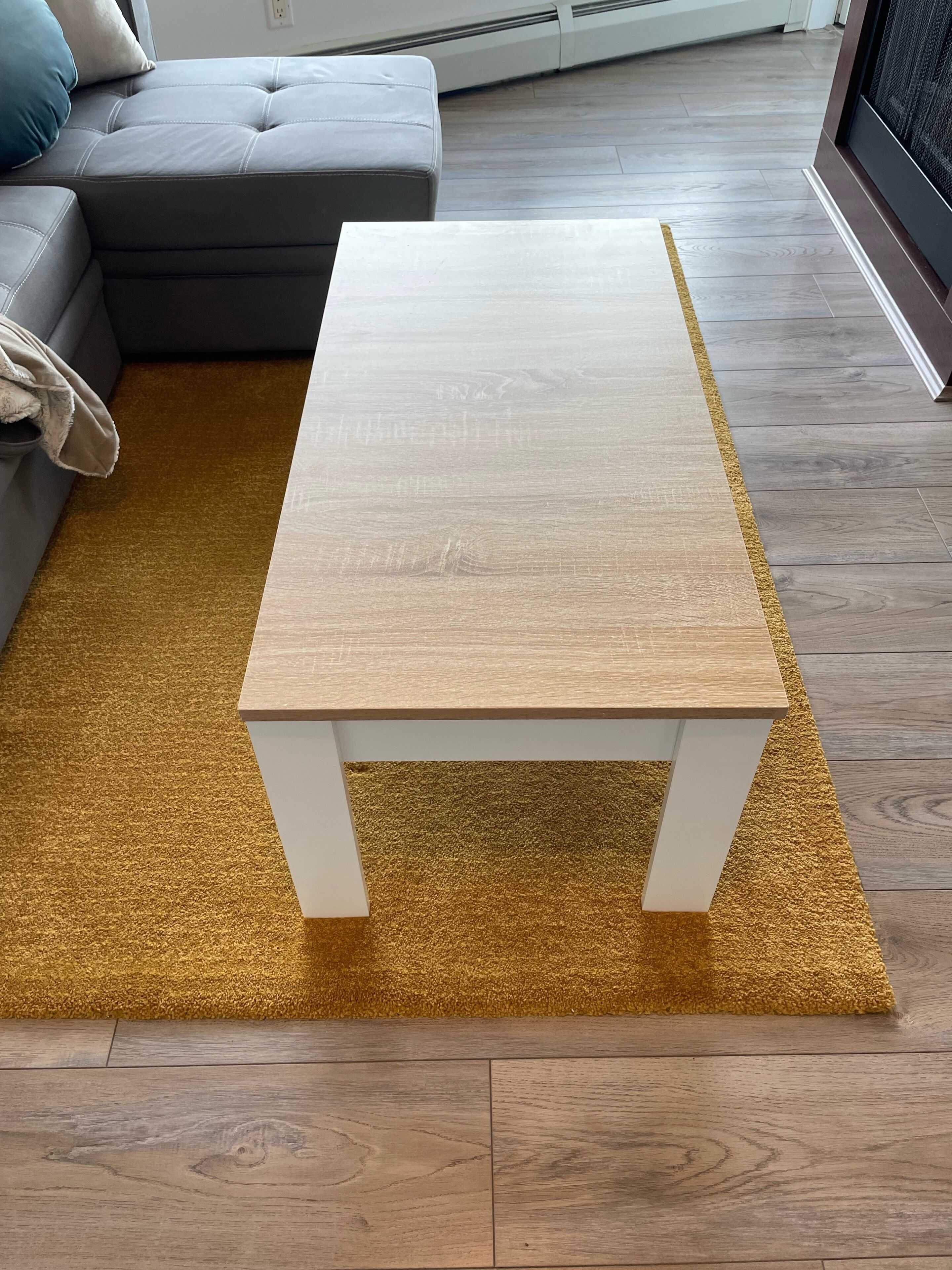A small white wooden table on a gold textured rug in a living room with a grey sofa and a white wall.