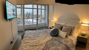 Een slaapkamer met een groot raam, een bed met beddengoed met een patroon, twee nachtlampjes en een aan de muur bevestigde televisie waarop een stadsgezicht te zien is.