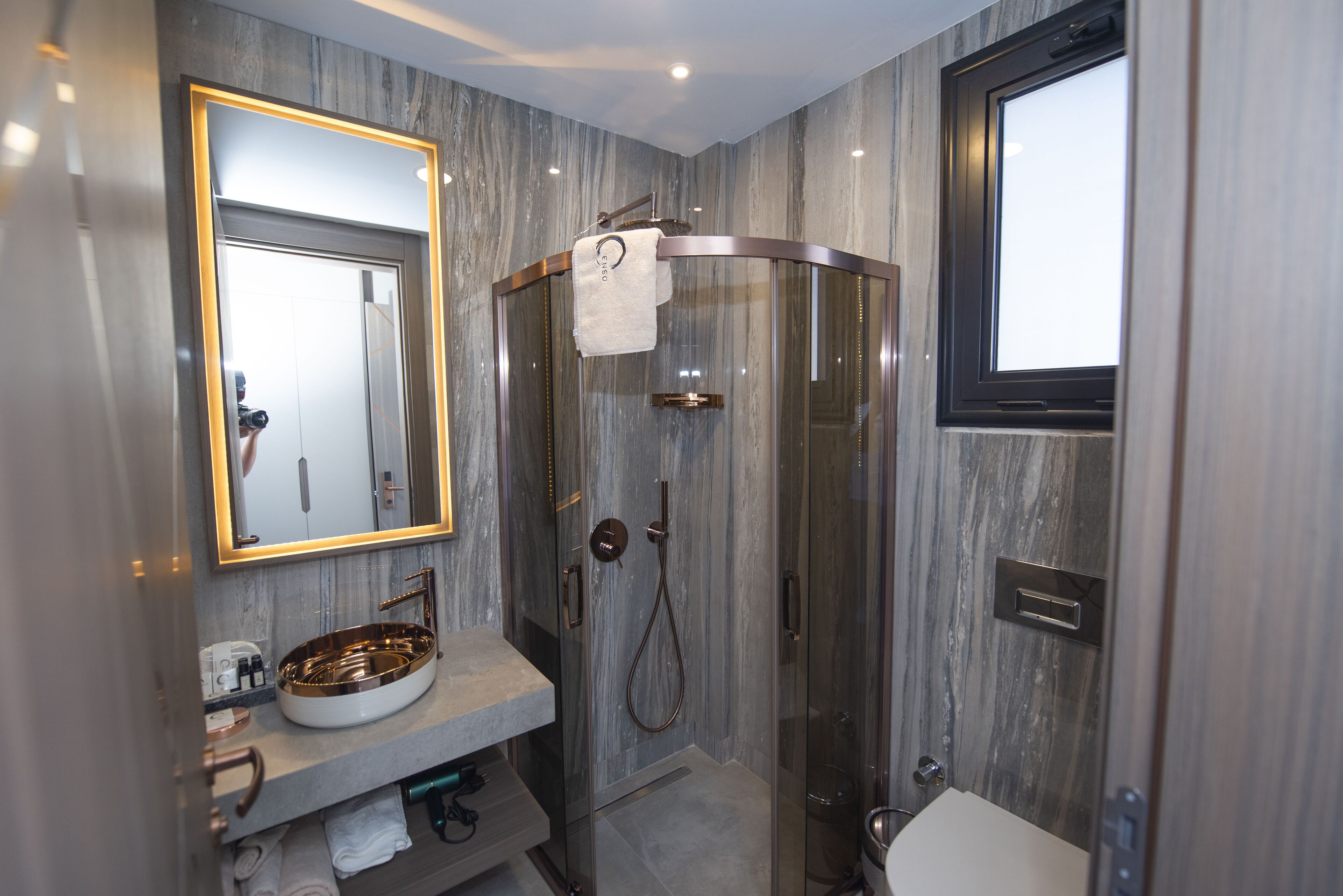 A modern bathroom with a curved glass shower, a marble wall, and a round sink with a gold faucet.