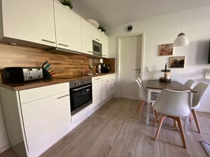 A modern kitchen with white cabinets, a wooden countertop, and a built-in oven. There is a dining area with white chairs and a table. - Haus Meeresblick Fewo A Stranddüne Ref 230902 - Haus Meeresblick Fewo A (Baabe)