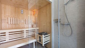 Een sauna met houten banken en een glazen wand, een douchegedeelte met een metalen douchekop, en een klein bankje met een opbergkist.