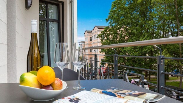 Een tafel met een schaal fruit, een wijnfles en glazen op een balkon met uitzicht op een stadsbeeld.