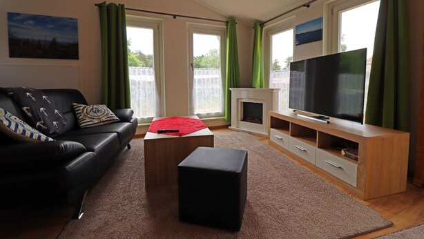 A living room with a black sofa, a red throw, a black ottoman, a TV on a wooden stand, and a fireplace. - House Zempin - Holiday homes Räuberkuhle 1 (Koserow)