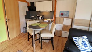 A compact kitchen with a dining table, chairs, and a small island. There is a black leather sofa with a striped pillow and a wooden cabinet with drawers. - House Zempin - Holiday homes Räuberkuhle 1 (Koserow)