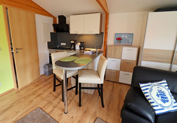 A compact kitchen with a dining table, chairs, and a small island. There is a black leather sofa with a striped pillow and a wooden cabinet with drawers.