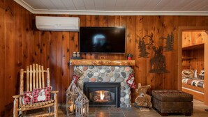A cozy living room with a stone fireplace, a television mounted on the wall, a rocking chair, and a wooden bench. - Koi Story Cabin - Lakefront, near Bike Trail (Maple Valley)