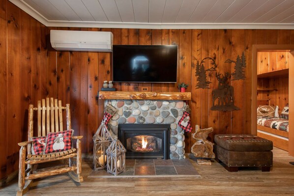 A cozy living room with a stone fireplace, a television mounted on the wall, a rocking chair, and a wooden bench.