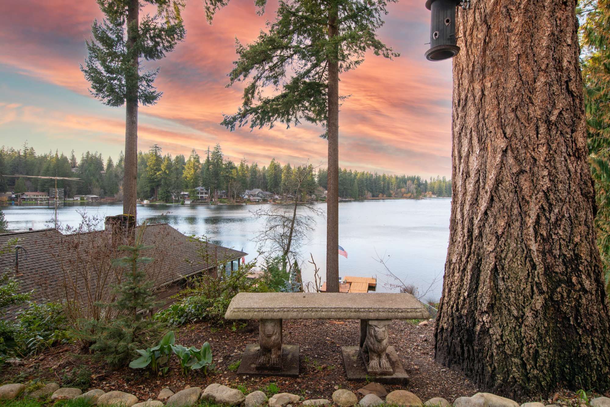 Un banc en pierre orné d’un drapeau, entouré de rochers et de végétation, surplombe un lac dont les rives sont bordées d’une forêt et où l’on aperçoit des maisons au loin.