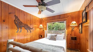 A wooden cabin bedroom with a bed, a ceiling fan, and a window with a view of greenery. - Koi Story Cabin - Lakefront, near Bike Trail (Maple Valley)