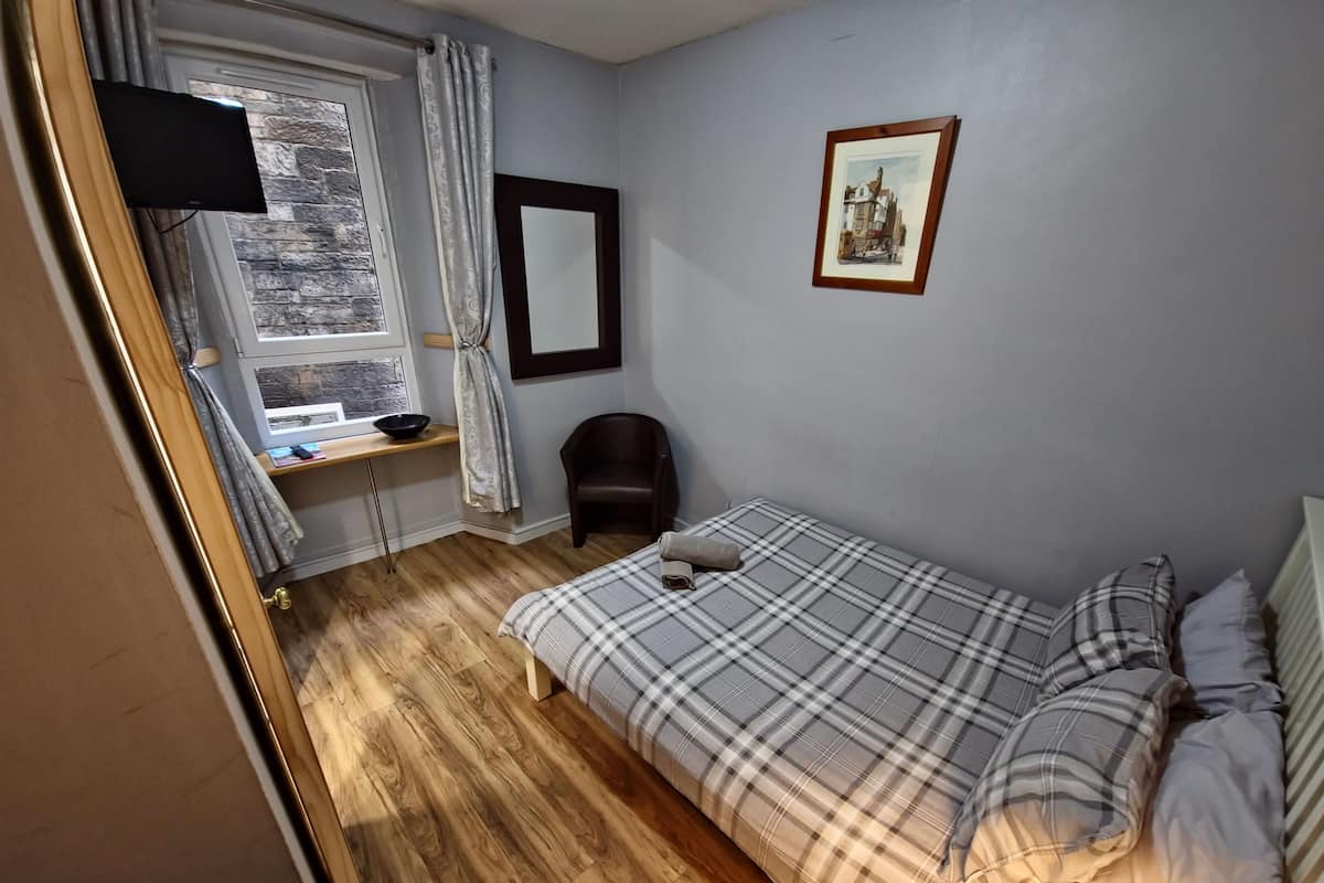 Ein Schlafzimmer mit Bett, Stuhl, Schreibtisch, einem Fenster mit Vorhängen, einem Spiegel und einem gerahmten Bild an der Wand.