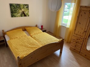 A bedroom with a wooden bed, a nightstand, a red lamp, a wooden wardrobe, a window with curtains, and a painting on the wall. - Large vacation apartment in an idyllic location with massage offer (Bisterschied)