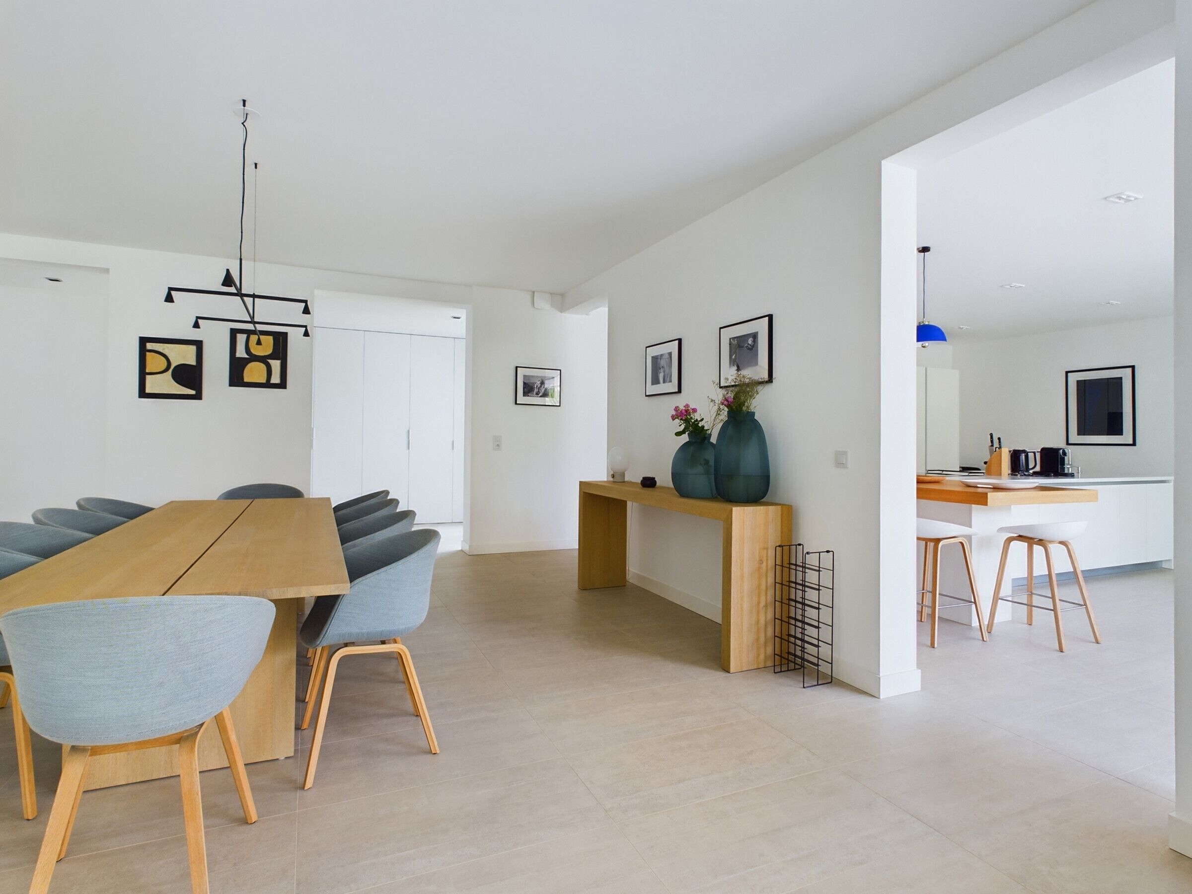 Un espace repas moderne avec une table en bois et des chaises grises, une cuisine en arrière-plan et un couloir menant à une porte.