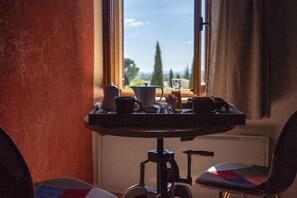 Une table avec une cafetière, des tasses et un verre de jus, placée près d’une fenêtre donnant sur des arbres et des bâtiments.