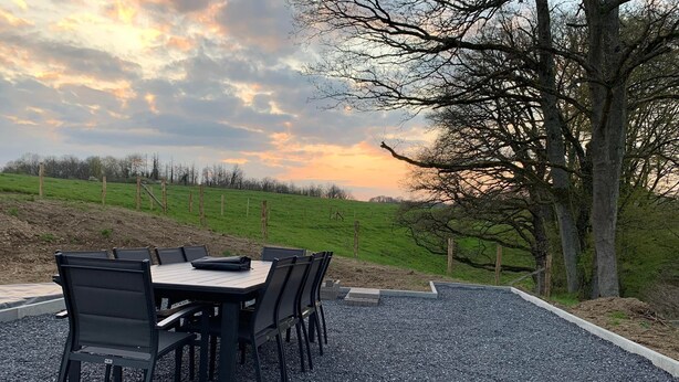 Un área de comedor al aire libre con una mesa y sillas, rodeada de grava, con vista a una colina cubierta de césped y árboles bajo un cielo nublado.