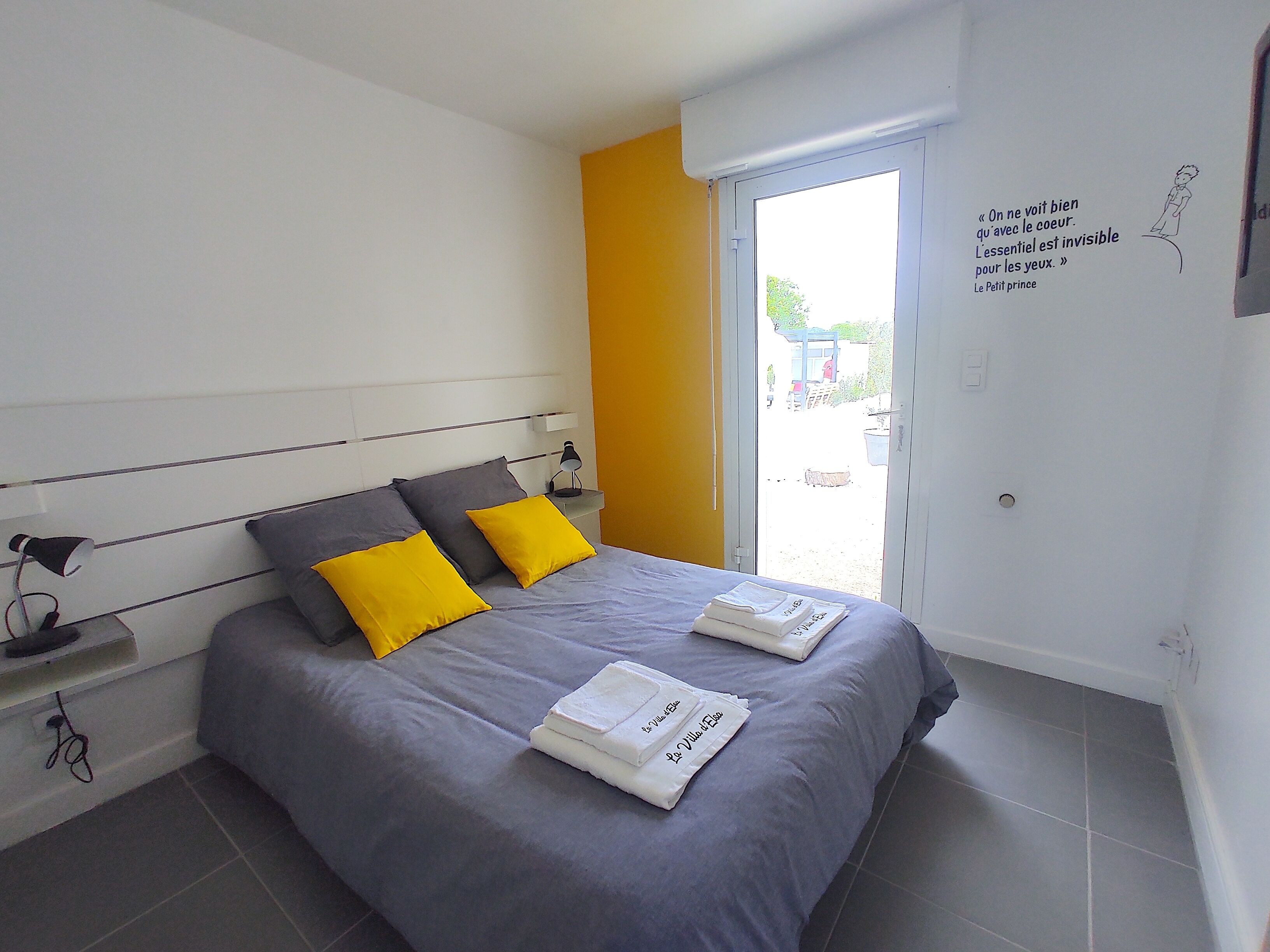 Een slaapkamer met een bed, kussens, een nachtkastje, een lamp en een muur met een citaat.