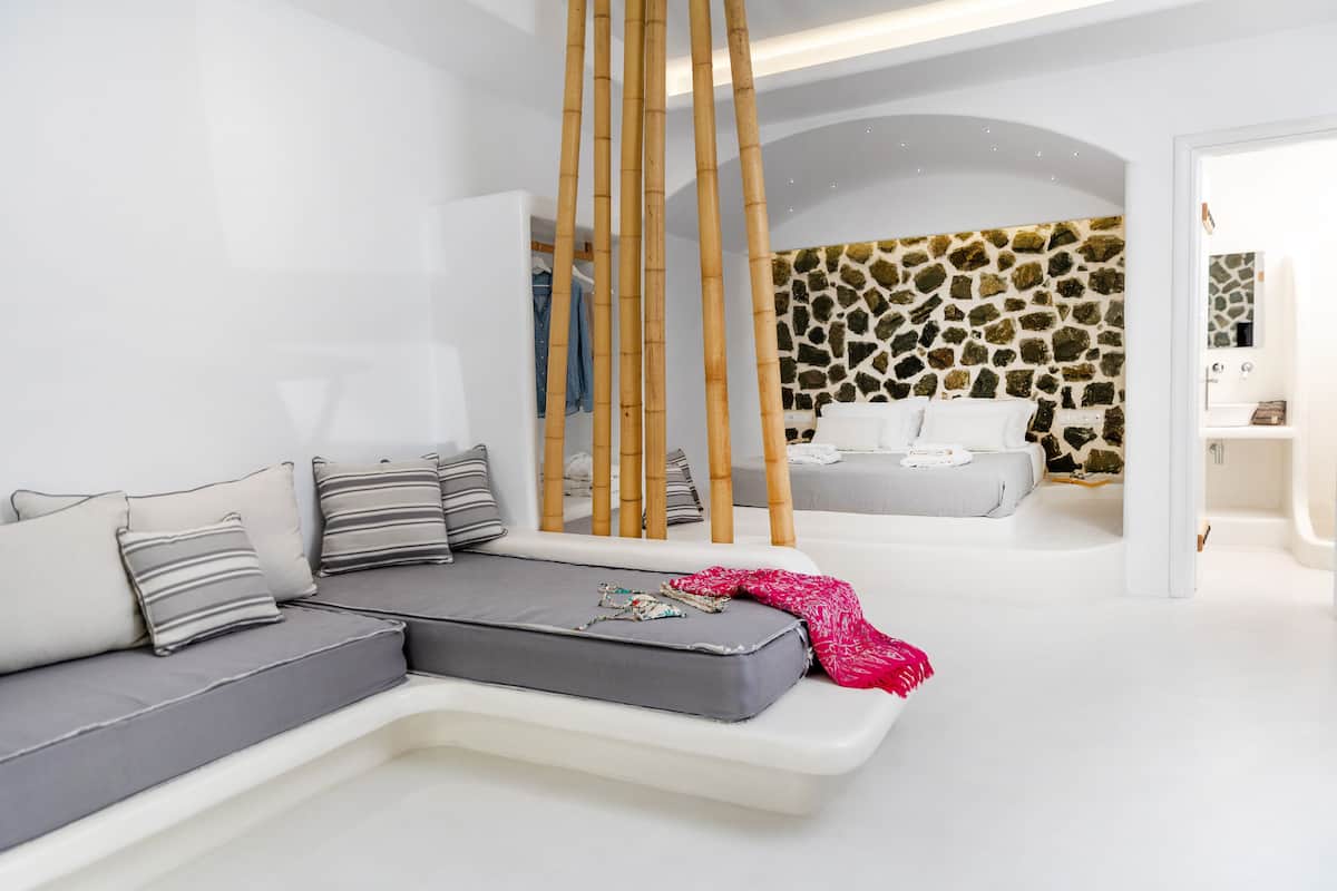 A modern, minimalist bedroom with a grey sofa, a bed with white linens, and a stone wall.