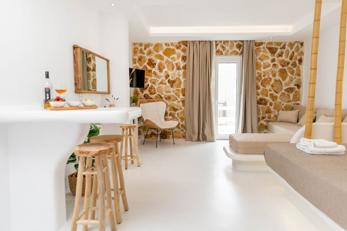 A modern hotel room with a stone wall, a sofa, a bed, a dining area with stools, and a large mirror.