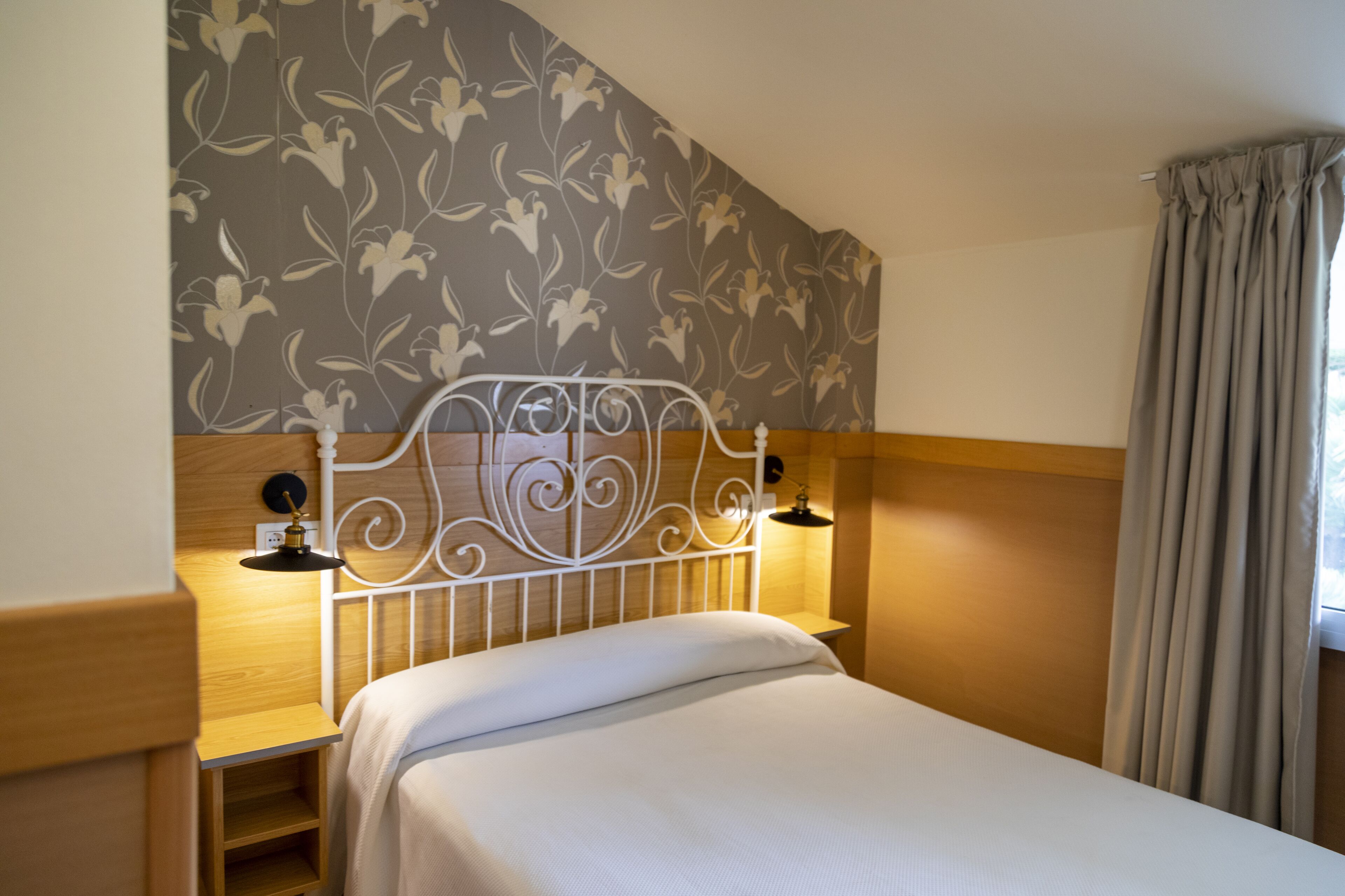A neatly made bed with a headboard, a bedside table with a lamp, and a floral-patterned wall.