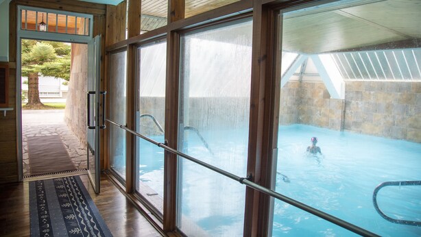 A swimming pool with clear glass walls, a wooden deck, and a stone wall in the background. - Hotel Ría de Suances (Suances)