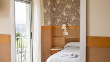 Habitación de hotel con cama, mesita de noche, lámpara de pared y vista a un balcón con barandilla.