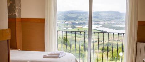 Habitación de hotel con cama, vista a un paisaje desde un balcón y ventana con cortinas.