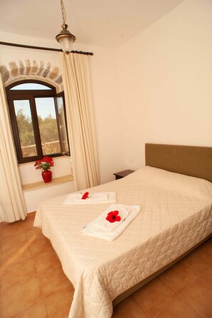 A hotel room with a large bed, a window with curtains, a vase with flowers, and a nightstand. - 3 bedroom villa common pool (Παλαιλώνι)