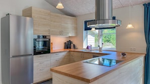Une cuisine moderne dotée d’un réfrigérateur en acier inoxydable, d’un four encastré, de meubles en bois, d’une grande fenêtre et d’un îlot central avec un plan de travail en verre.