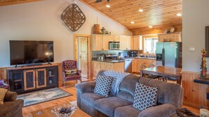 A cozy living room with a large TV, a leather armchair, and a sofa with patterned cushions. The kitchen features wooden cabinets, a microwave, and a refrigerator. - Log Cabin in West Jefferson - Fishing Pong + Hot Tub + Fire Pit + Mountain Views (West Jefferson)