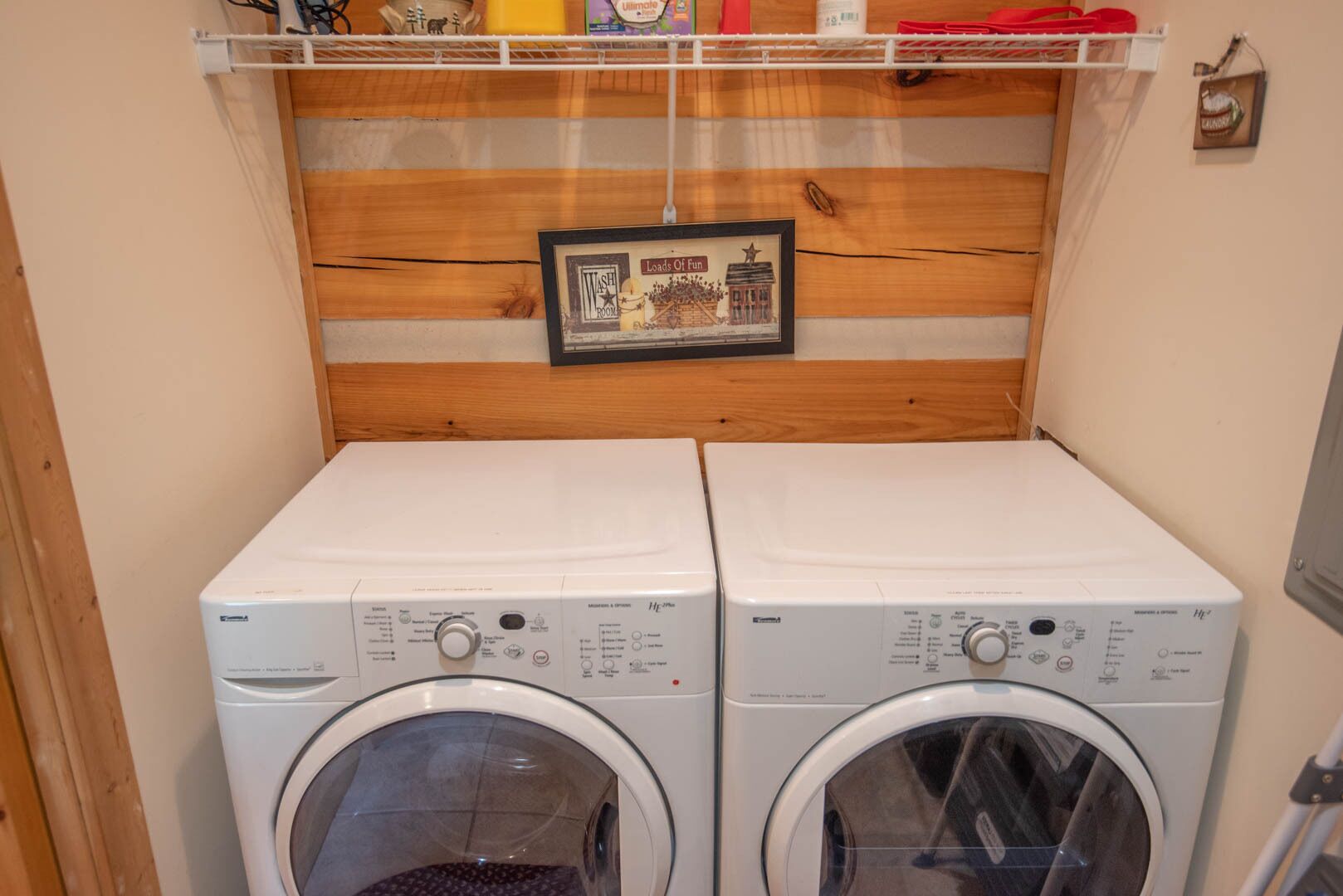 A laundry room with two front-loading washing machines and a wooden shelf above them.