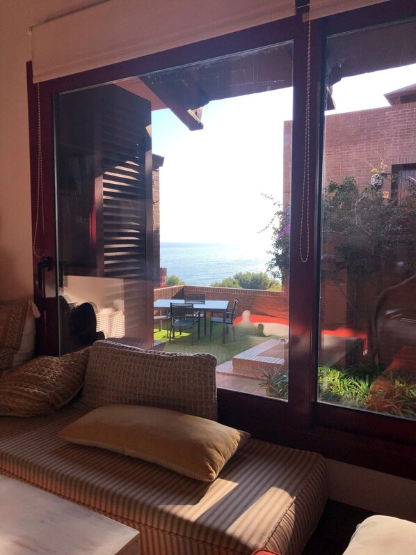 A room with a large sliding glass door leading to a patio with a view of the sea. There is a sofa with cushions and a small table with chairs.