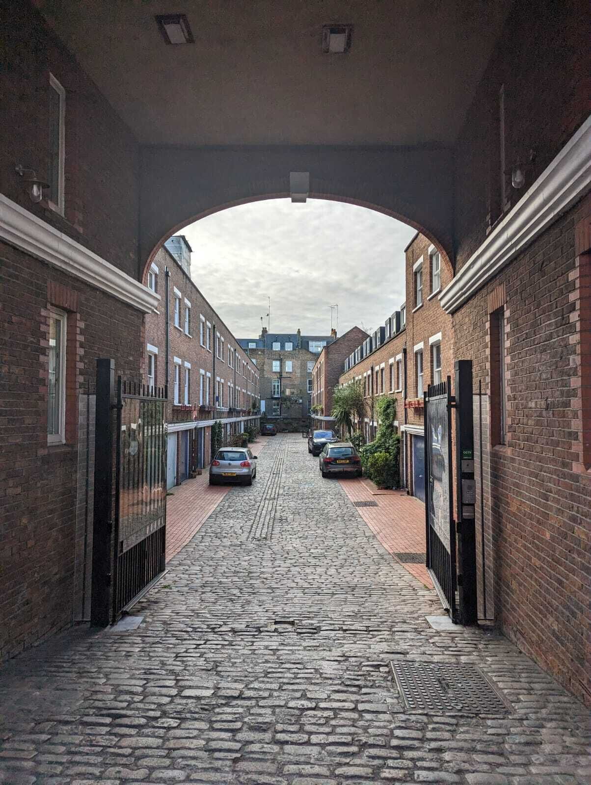 A cobblestone street with brick buildings, a covered archway, and parked cars.